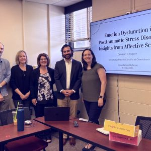 Photo of Cameron Pugach & his committee next to his dissertation title slide