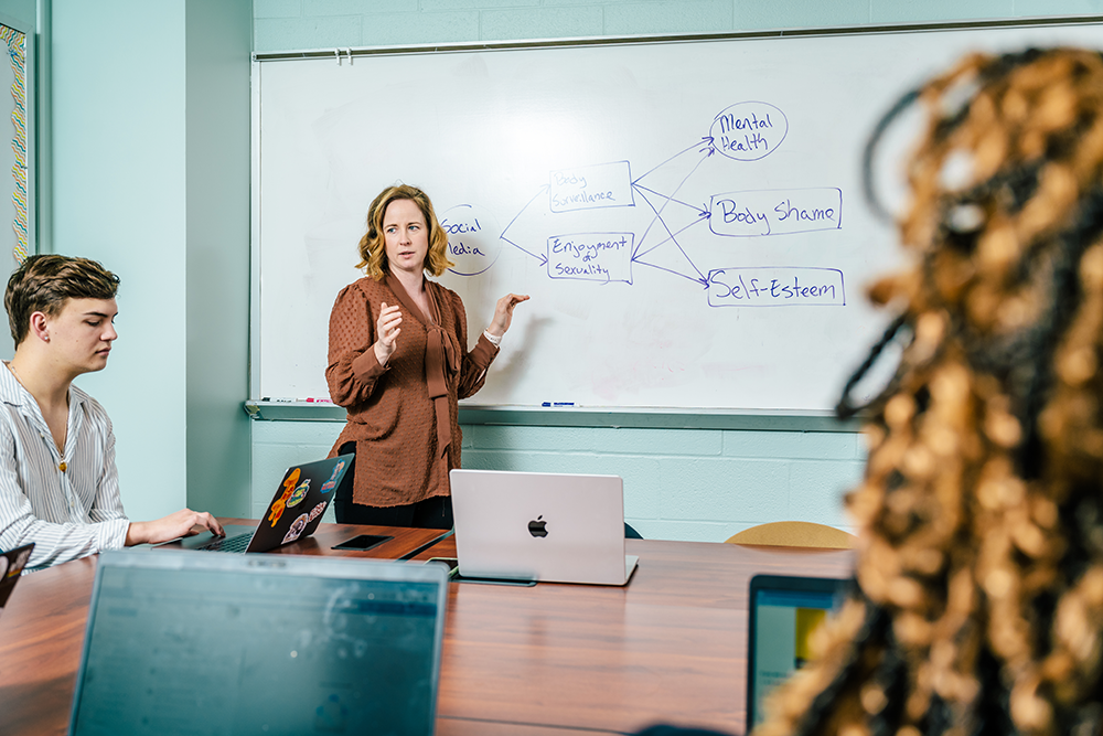 Psychology professor teaching to two students with social media and self esteem information on the white board