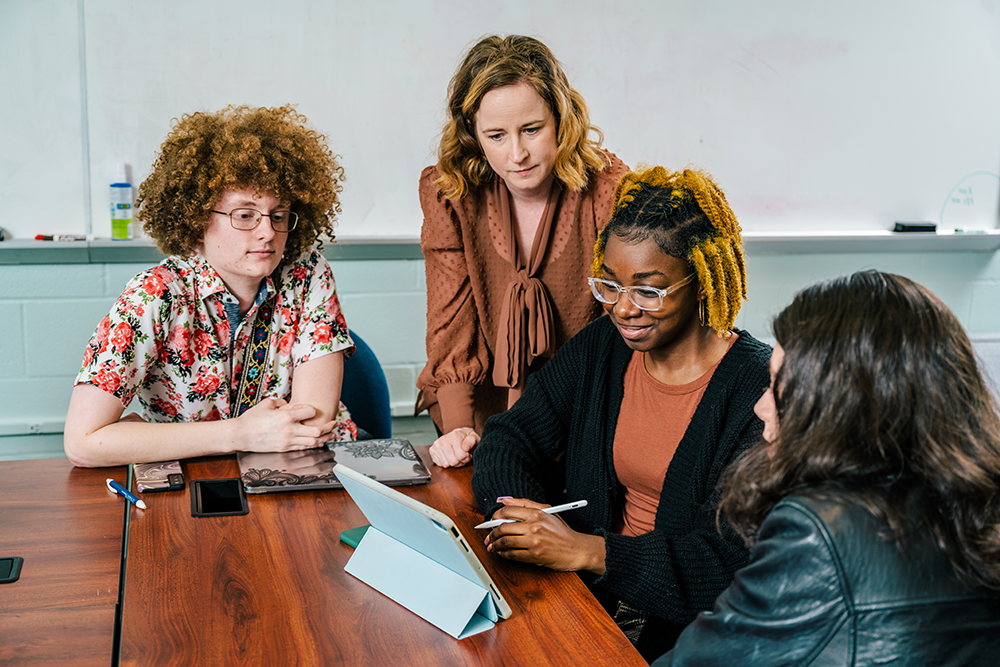 Three undergraduate students work together at table with Professor Jensen looking over shoulders